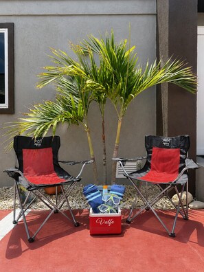 Zwei Gartenstühle mit roten Kissen, ein kleiner Baum und ein roter Kühlbox mit dem Schriftzug 'Valfe' auf roter Fläche.