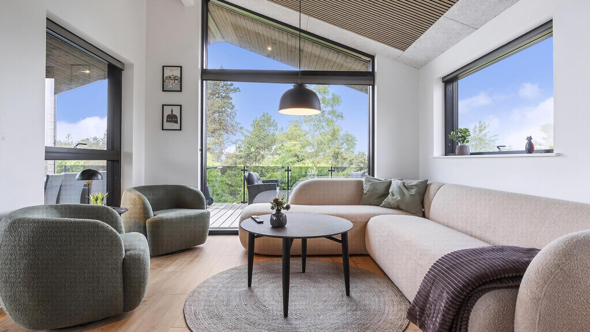 A modern living room with a sofa, armchairs, a coffee table, and large windows offering a view of trees.