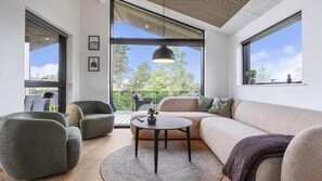 Ein modernes Wohnzimmer mit einer Couch, Sesseln, einem Couchtisch und großen Fenstern mit Blick auf Bäume.