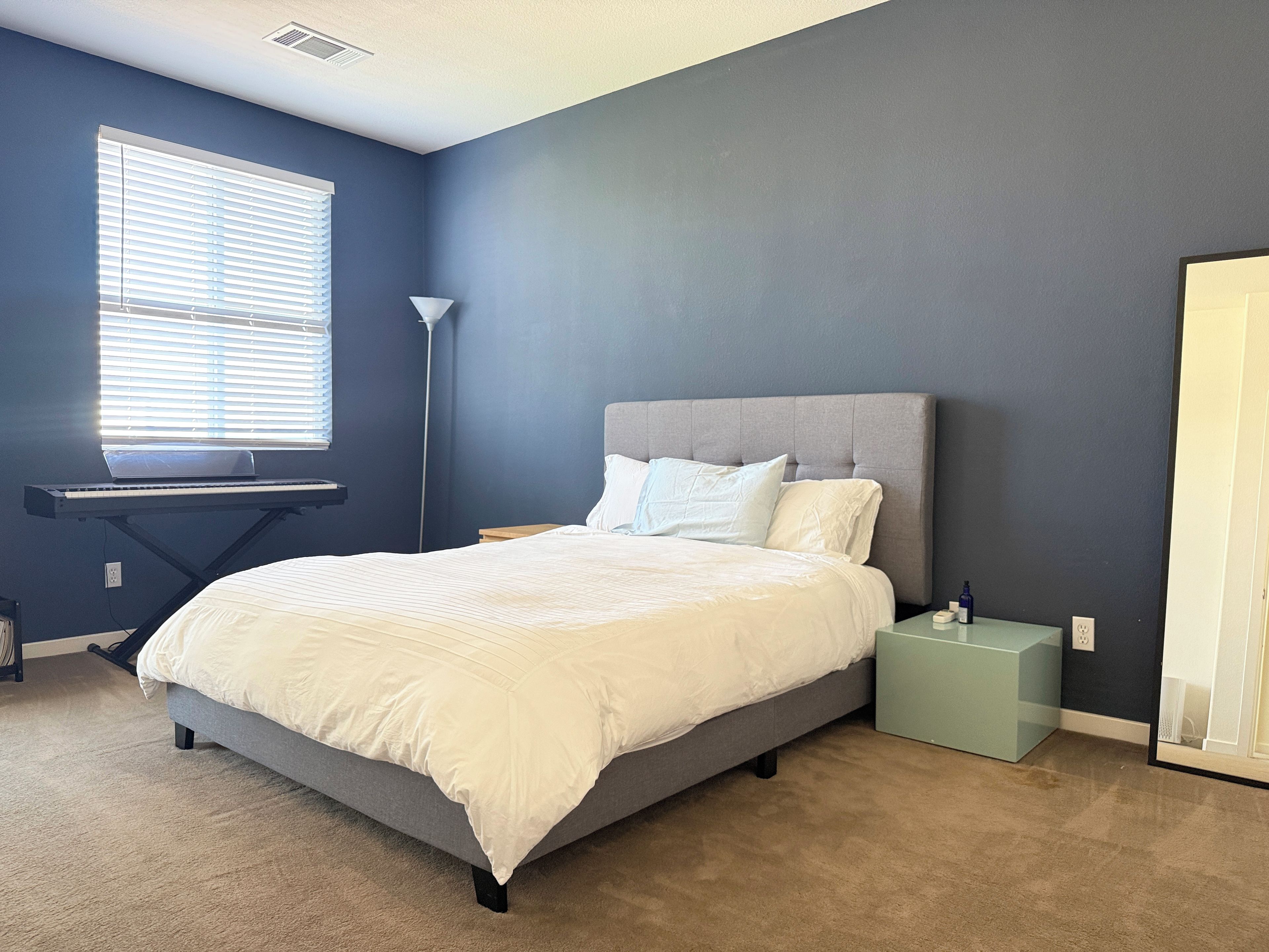 A bedroom with a bed, a standing lamp, a window with blinds, and a desk with a laptop.