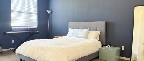 A bedroom with a bed, a standing lamp, a window with blinds, and a desk with a laptop.