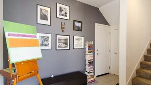 Een gang met een schilderij op een lessenaar, een stapel boeken, ingekaderde foto's aan de muur en een deur.