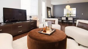 Een moderne woonkamer met een flatscreen televisie, een bruin poefje, witte banken, een eethoek met een tafel en stoelen, en een grote spiegel.