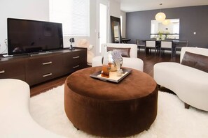 A modern living room with a flat-screen TV, a brown ottoman, white sofas, a dining area with a table and chairs, and a large mirror. - Modern LUXE 2B-2.5B townhome near downtown San Jose with AC and Peloton Bike (San Jose)