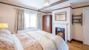 A bedroom with a bed, a fireplace, a bookshelf, and a window with curtains. - Colston Cottage, Badingham (Badingham)
