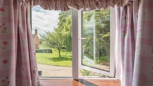 A room with a large sliding glass door leading to a garden, a wooden floor, and a window with a view of greenery and a building. - Colston Cottage, Badingham (Badingham)