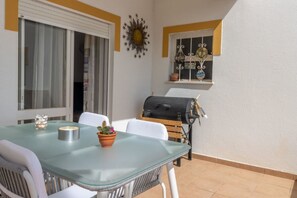 Eine Terrasse mit Glastisch, weißen Stühlen, einem Grill und einem Fenster mit Dekoration.