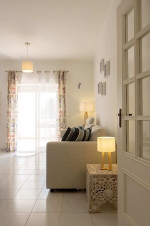 Ein Schlafzimmer mit Bett, Nachttisch mit Lampe, einer Tür mit Glasfenstern und Blick ins Freie durch ein Fenster mit Vorhängen.