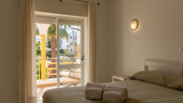 Un dormitorio con una cama, una mesita de noche, una lámpara de pared y un ventanal corredero que da a un espacio exterior con una silla y palmeras.