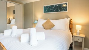 A neatly made bed with white linens and towels, a bedside lamp, a mirror, and a wall-mounted artwork. - Barnaby Cottage, Southwold (Southwold)