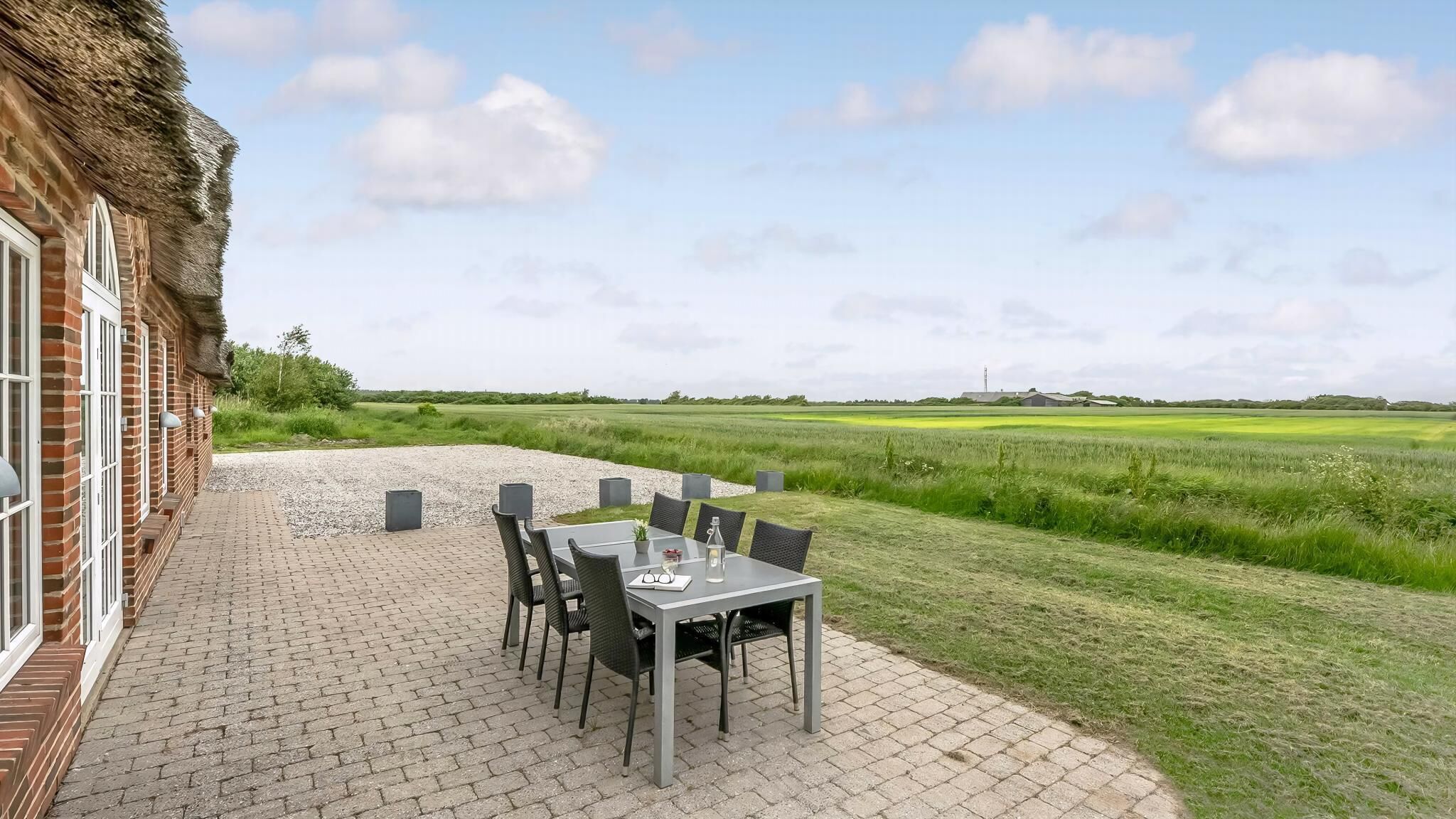 A patio with a table and chairs, a thatched-roof building, and a view of a green field.