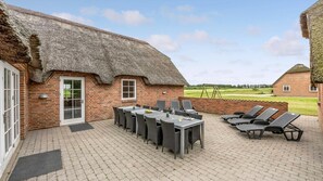 A thatched-roof building with a patio area featuring a table and chairs, a swing set, and a view of a golf course. - VillaVilla 982 - Vedersø Klit, Vestjylland (Ulfborg)