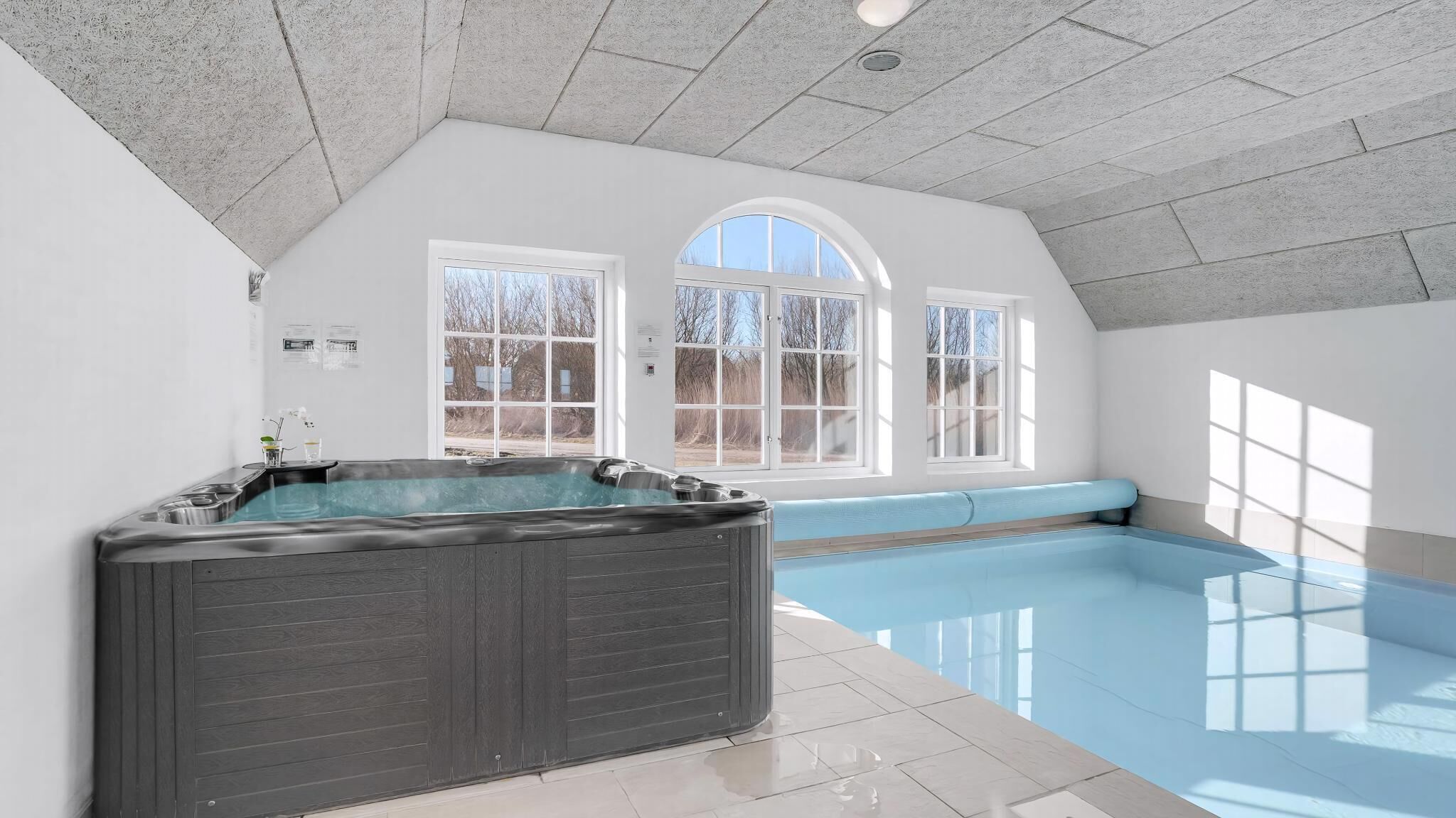 A spacious indoor swimming pool with a hot tub, large windows, and a high ceiling.