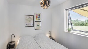 A bedroom with a bed, a window with a view of greenery, and two wall-mounted artworks. - VillaVilla 969 - Marielyst, Lolland/Falster/møn (Væggerløse)