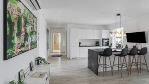 A modern kitchen with white cabinets, a central island with bar stools, a flat-screen TV mounted above the island, and a large abstract painting on the wall. - VillaVilla 969 - Marielyst, Lolland/Falster/møn (Væggerløse)