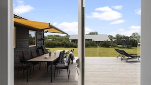 Eine Holzterrasse mit Sitzgelegenheiten im Freien, ein gelbes Markisenvorzelt und Blick auf eine Rasenfläche mit einem Haus und einem Trampolin.