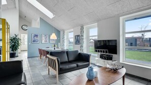 Una sala de estar moderna con un sofá de cuero negro, una mesa de centro de madera y un televisor montado en la pared. Hay una zona de comedor con una mesa y sillas, y un gran ventanal que ofrece vistas al exterior.