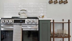 Eine Küche mit einem Edelstahlherd, einem Backofen und einem Wasserkocher. Ein Holzregal zeigt Gläser, und ein Metallregal beherbergt Tassen.