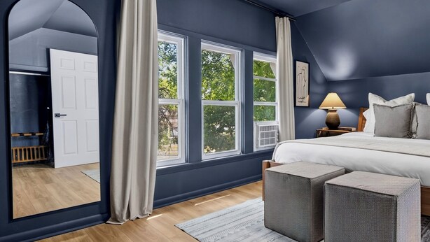 Ein Schlafzimmer mit einem großen Bett, zwei Nachttischen, einer Lampe und Blick auf Bäume durch die Fenster.