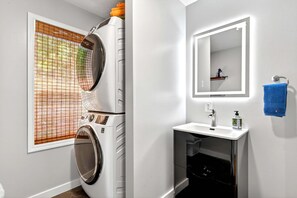 Ein Waschraum mit gestapelten Waschmaschinen und Trocknern, ein Fenster mit Jalousien, ein Waschbecken mit Spiegel und ein blaues Handtuch an einem Wäscheständer.