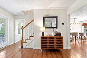 A staircase with wooden balusters, a wooden console table with a mirror above it, and a dining area with chairs and a table in the background. - Calm Water Cove | Private Dock on Lake James, Game Room, Fire Pit & Spa Suite — Dogs Welcome! (Nebo)