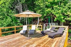 A wooden deck with lounge chairs and a large umbrella, surrounded by lush greenery. - Calm Water Cove | Private Dock on Lake James, Game Room, Fire Pit & Spa Suite — Dogs Welcome! (Nebo)