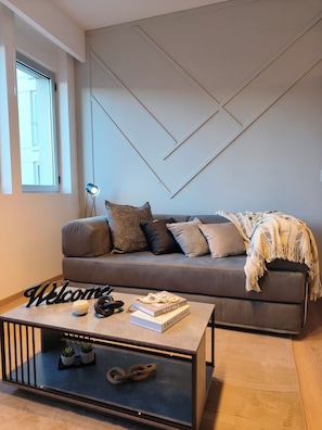 Een moderne woonkamer met een grijze bank, een bijzettafeltje met boeken en een welkomstbord, en een geometrisch wandontwerp.