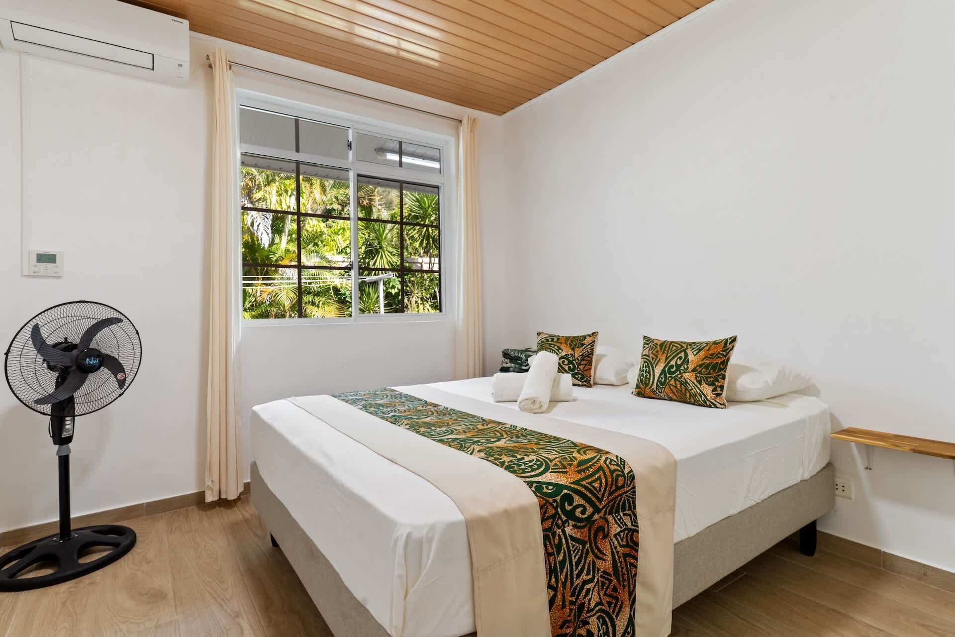 A bedroom with a bed, a standing fan, a window with a view of greenery, and a wooden ceiling.