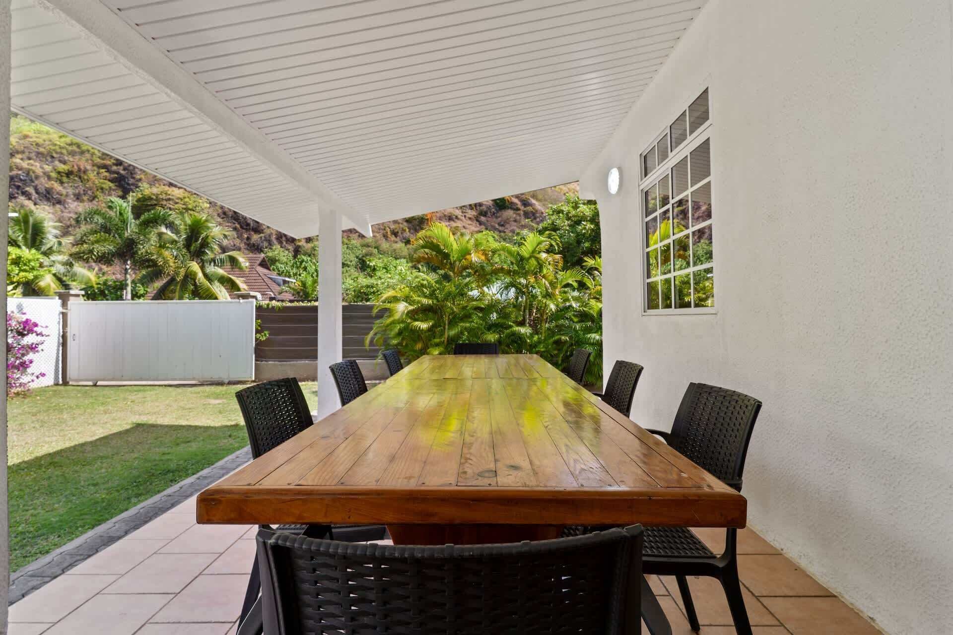 A covered patio with a wooden table and black chairs, surrounded by white walls and a view of greenery.