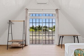 A room with a bed, a small table, a hanging chair, and a balcony with a view of palm trees.
