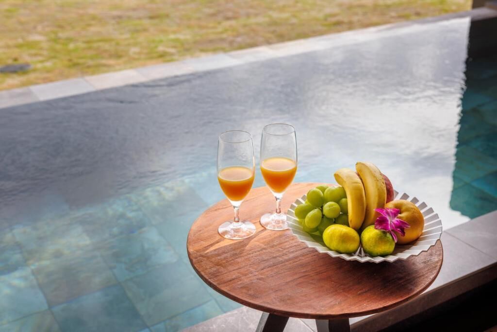 A round wooden table with two glasses of orange juice and a plate of fruit by a pool.
