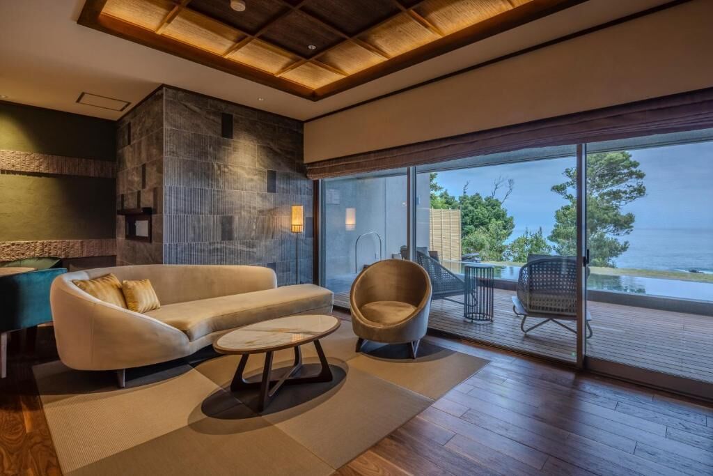 A modern living room with a sofa, armchair, and coffee table, a stone accent wall, and a large sliding glass door leading to a balcony with a view of the sea.