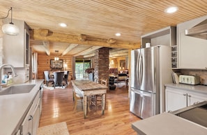 A rustic kitchen with wooden beams, a stone fireplace, a dining area with a wooden table, and a modern refrigerator. - Historic Cowboy Hall of Fame Ranch (Condon)