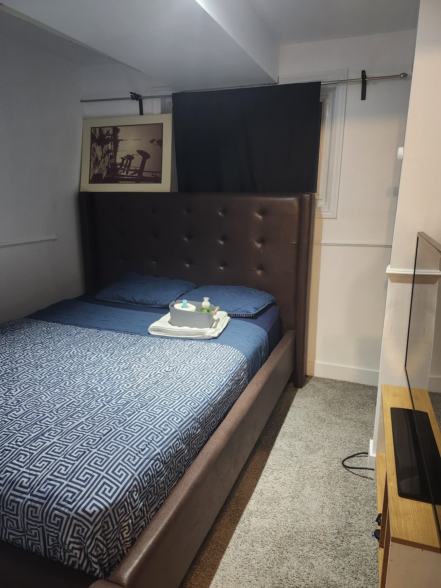 Un dormitorio con cama, una mesita de noche, televisor montado en la pared y un cuadro enmarco.