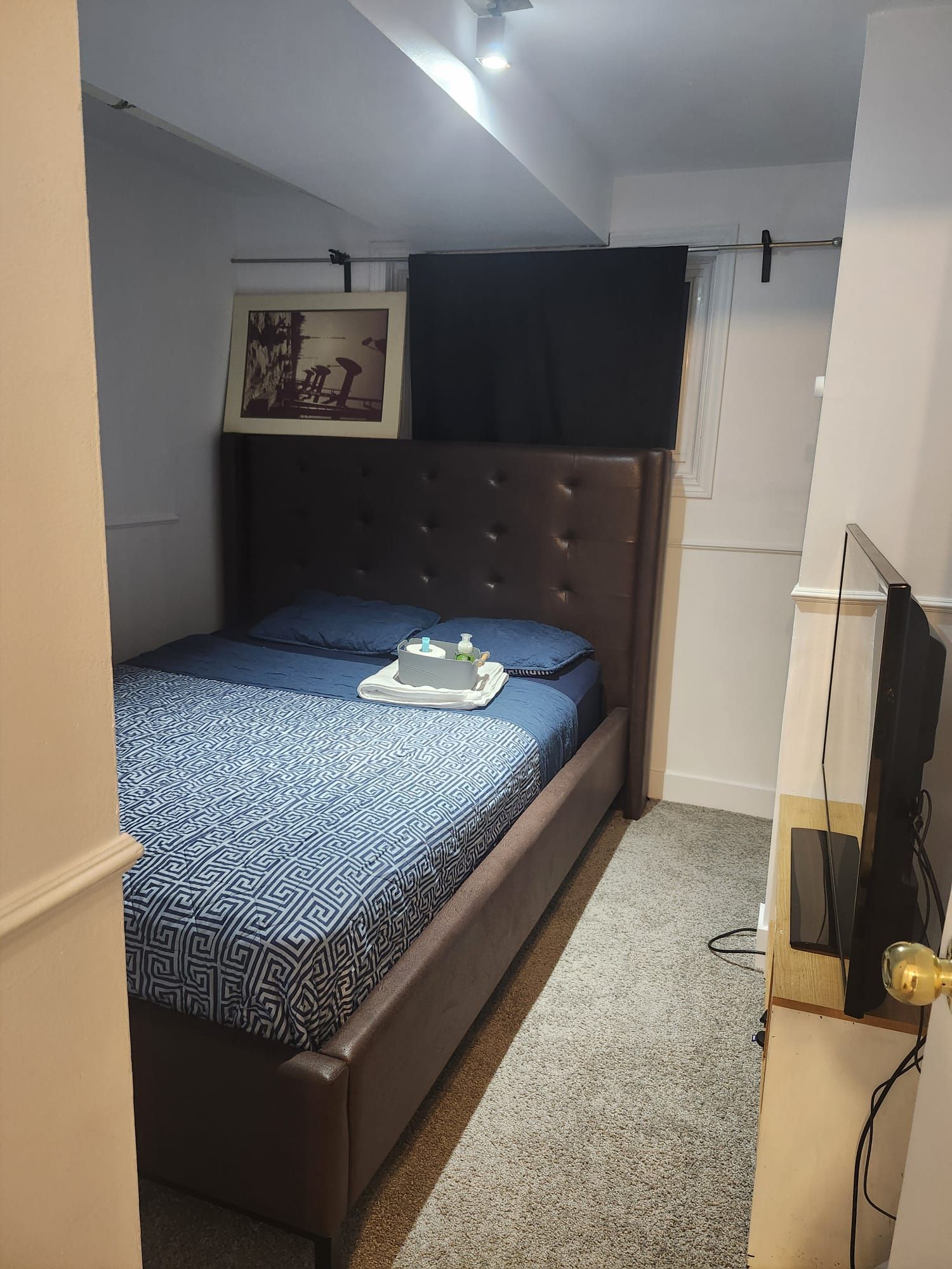 Une petite chambre à coucher, bien rangée, avec un lit, un tableau encadré au mur et un téléviseur posé sur un meuble.