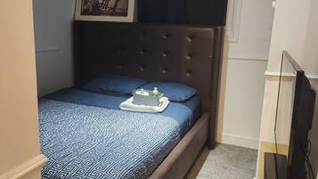 Une petite chambre à coucher, bien rangée, avec un lit, un tableau encadré au mur et un téléviseur posé sur un meuble.