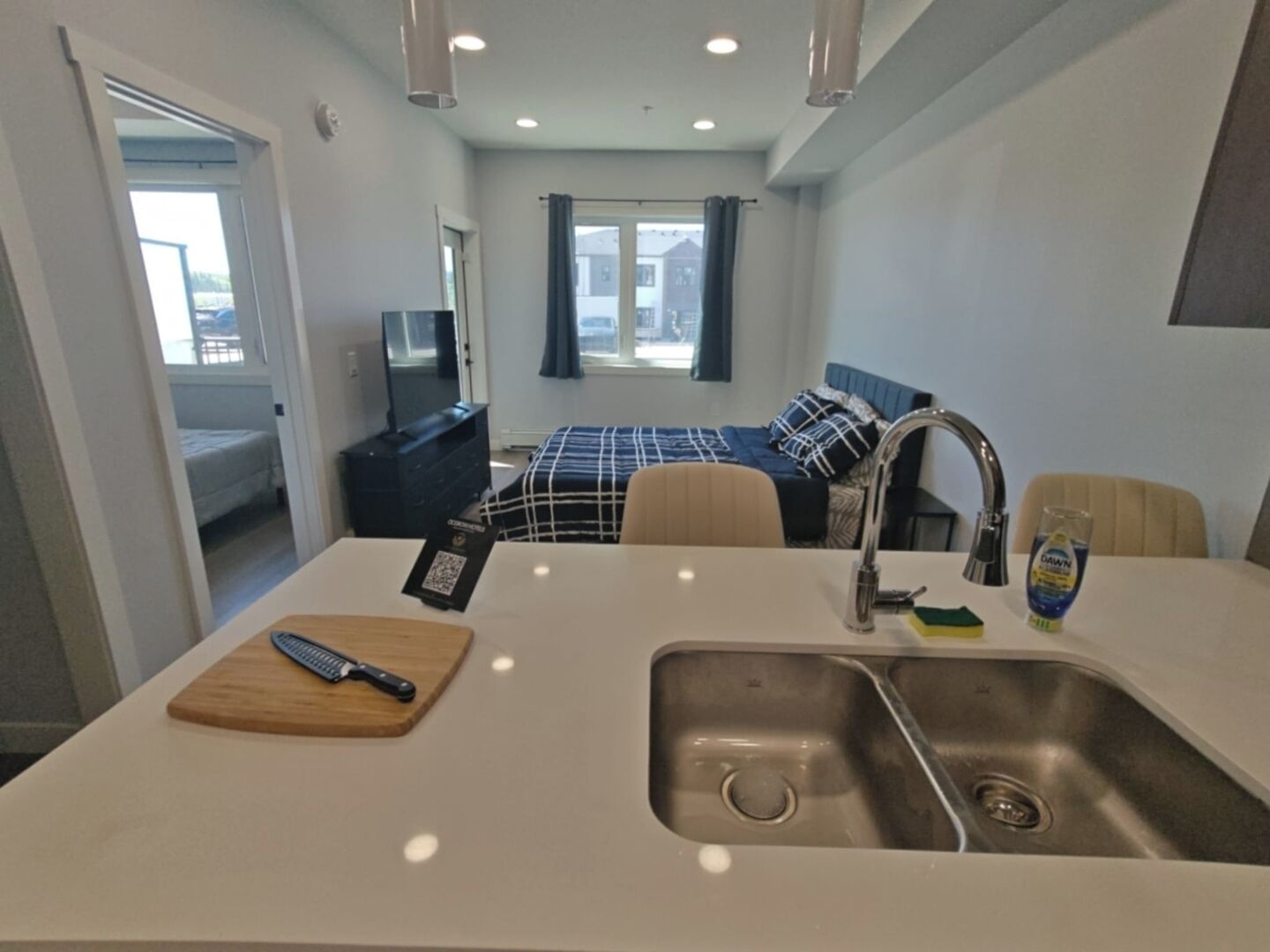 A compact hotel room with a kitchenette, a bed with a blue headboard, a television, and a window with curtains.