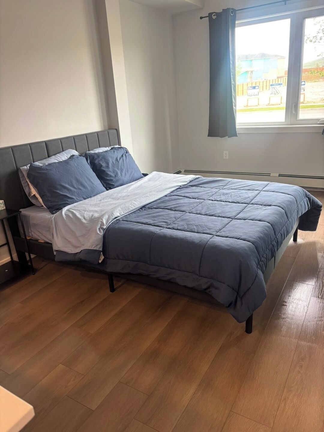 A bedroom with a bed, a nightstand, a window with curtains, and a view of the outside.