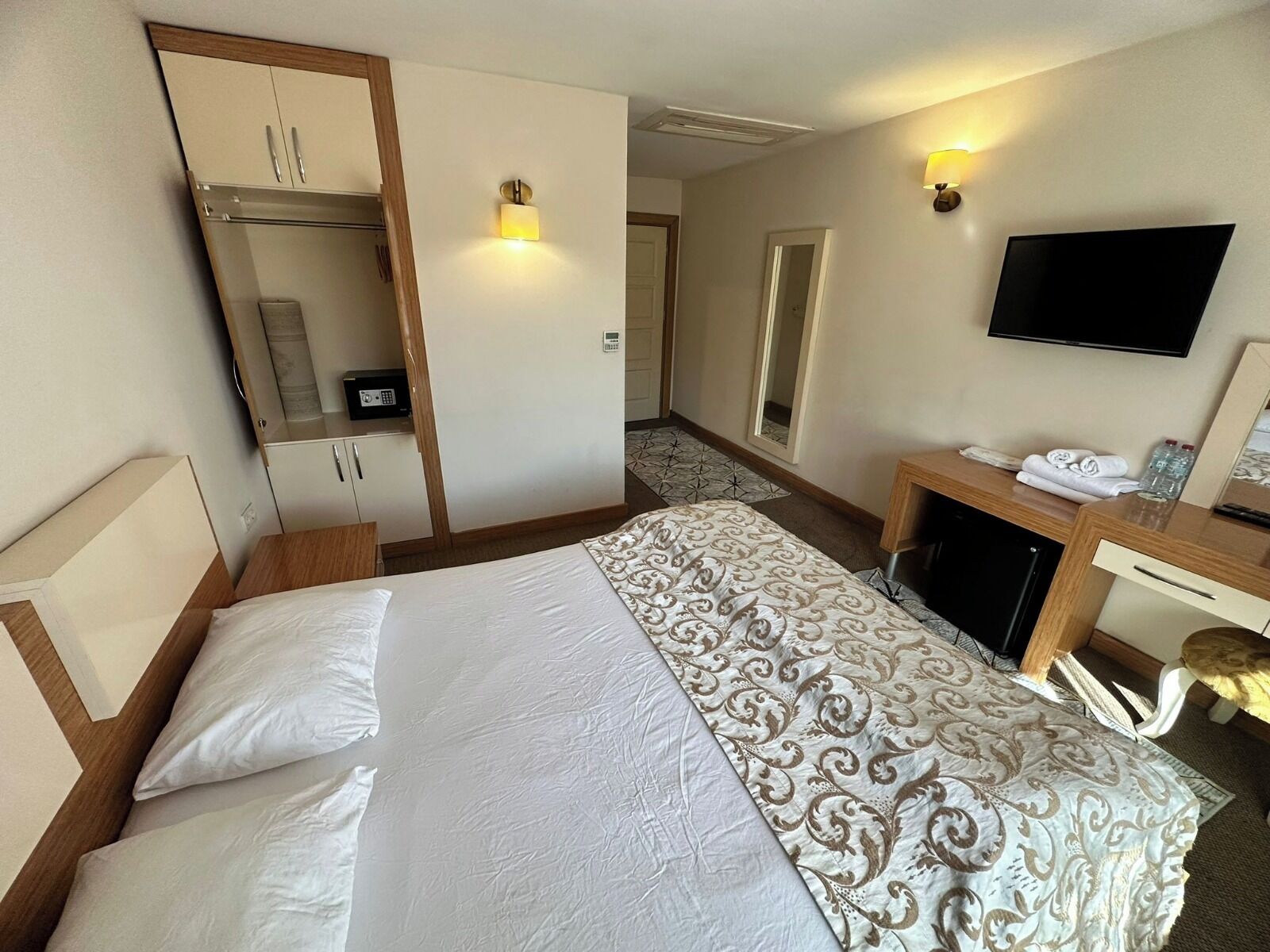 A hotel room with a bed, a desk with a chair, a TV, a mirror, and a wardrobe.