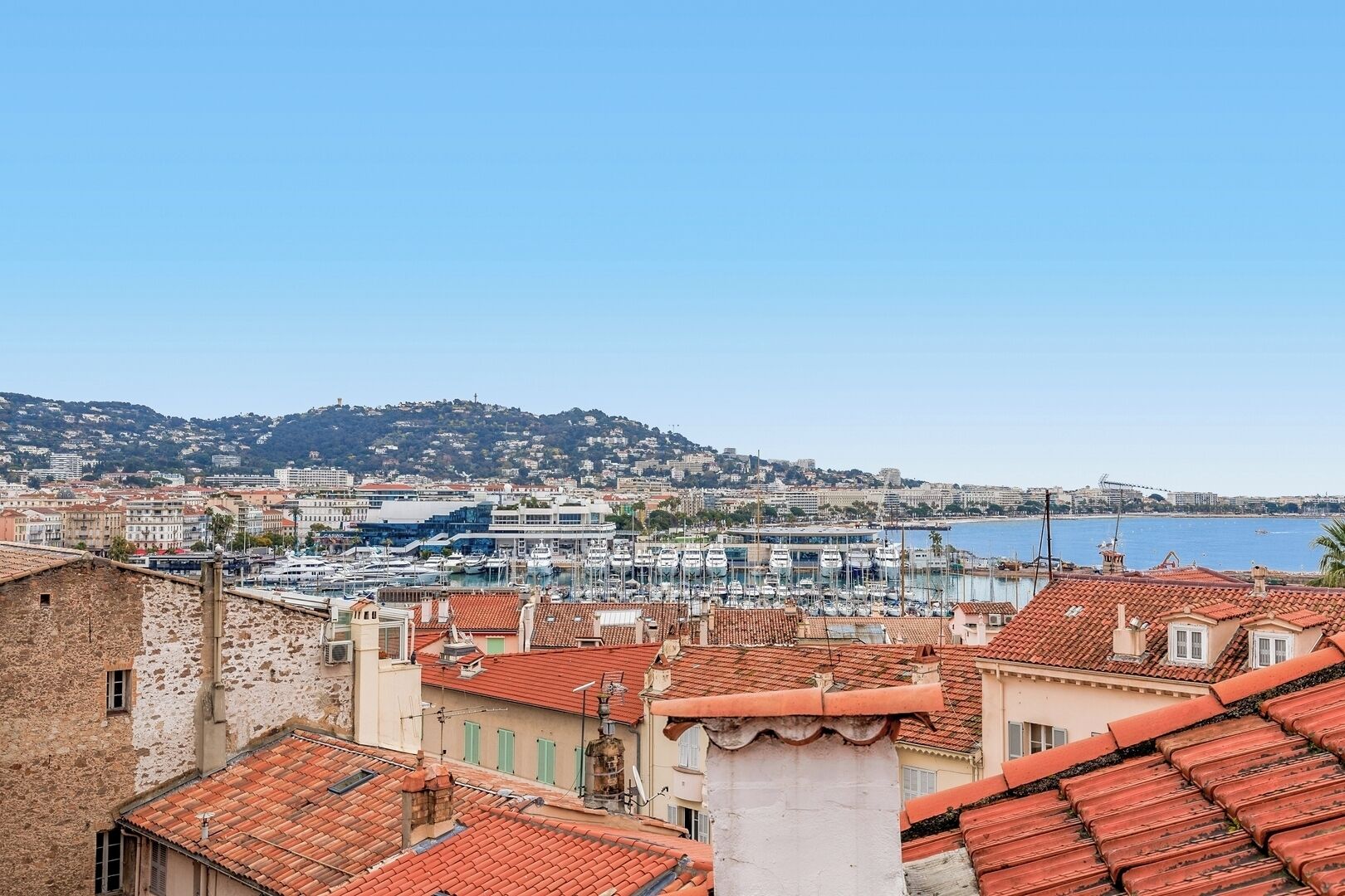 A coastal cityscape with a marina, terracotta-roofed buildings, and a hilly backdrop.