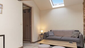 Ein kleiner, gemütlicher Raum mit einer Couch, einem Tisch und zwei Hocker. Eine Tür führt in einen anderen Raum, über der Couch ein Oberlicht.