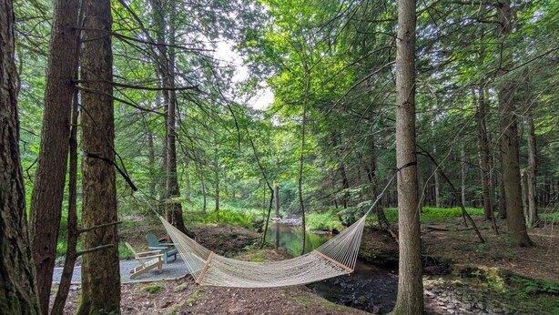 A hammock in a forest with a stream and a chair. - Charming 3-bedroom chalet in marvelous Livingston Manor with a majestic Stream (Livingston Manor)