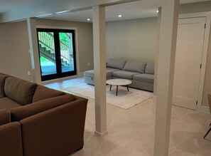 A living room with a brown sofa, a grey sofa, and a white coffee table. There is a sliding glass door leading to a balcony with a view of a staircase and greenery.