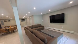 A modern living room with a sectional sofa, a dining table with chairs, and a flat-screen TV mounted on the wall. - Charming 3-bedroom chalet in marvelous Livingston Manor with a majestic Stream (Livingston Manor)