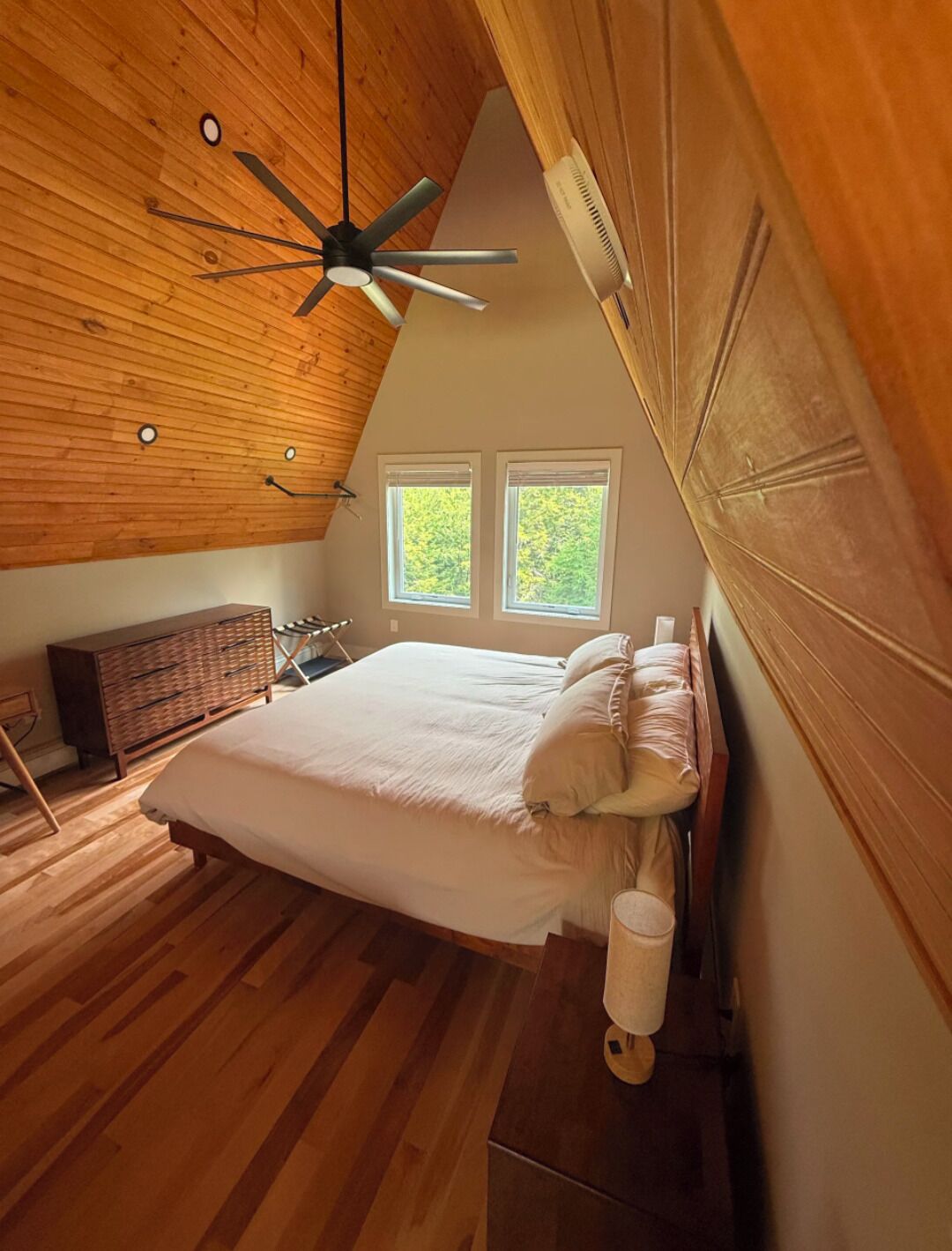 Une chambre avec un plafond en pente, un lit, une commode et deux fenêtres.