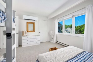 Ein Schlafzimmer mit einem Bett, einem Schminktisch, einem Spiegel, einem Standventilator und einem Fenster mit Blick auf Häuser.
