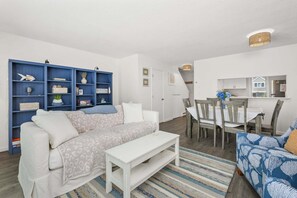 Ein Wohnzimmer mit einer Couch, einem weißen Couchtisch, einem blauen Bücherregal, einem Esstisch mit Stühlen und einer Küchenzeile im Hintergrund.