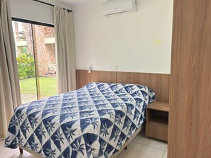 Ein Schlafzimmer mit einem Bett, einem Nachttisch aus Holz, einem Fenster mit Vorhängen und einer Klimaanlage.