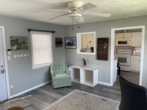 A room with a ceiling fan, a light switch, a door, a window with blinds, a chair, a small table with a decorative bird, wall-mounted artwork, and a kitchen area with cabinets and appliances.