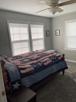 A bedroom with a bed, two windows with blinds, a ceiling fan, and a carpeted floor.